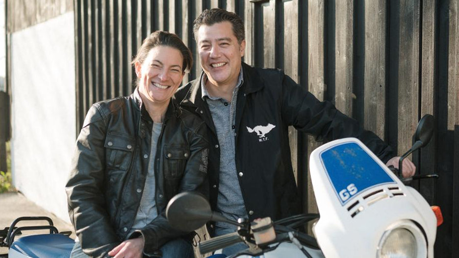 Two people sitting on a motorcycle with a wooden fence in the background